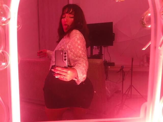HairyValeri with medium-length brown hair, white patterned shirt, and black skirt, showing her ass in a mirror selfie, red lighting, tongue out, holding a silver phone.