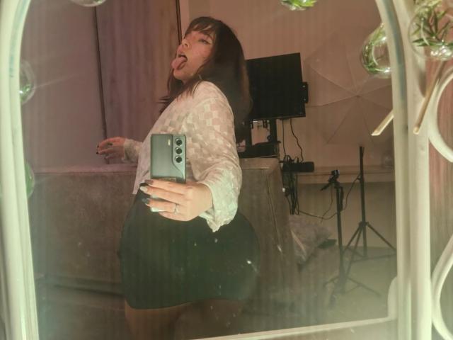 HairyValeri with dark brown hair, tongue out, wearing a white patterned shirt and black skirt, taking a mirror selfie with a phone, showing her round ass.