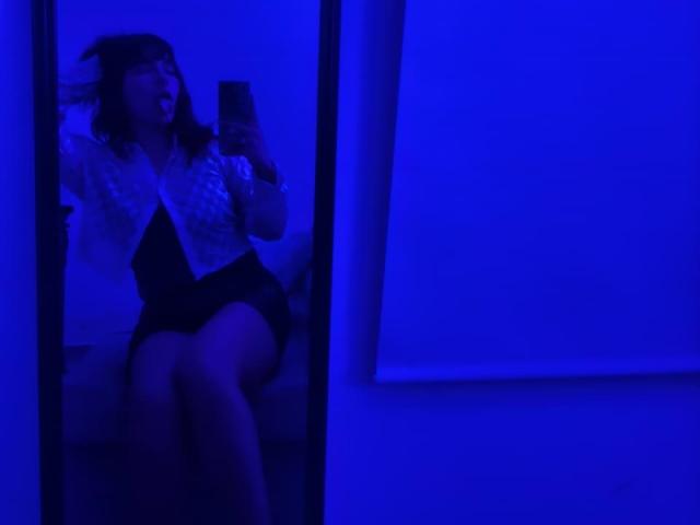 HairyValeri with blue-tinted skin, wearing a white lace shirt and black skirt, sitting, holding a phone, legs crossed, in a mirror selfie, showing her ass.