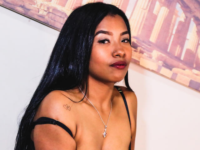 SophiaMullerr mit langem, schwarzem Haar, brauner Haut, rotem Lippenstift, einem kleinen Tattoo an der rechten Schulter, einer Halskette, einem schwarzen Trägerkleid, das ihre Brüste zeigt, und die in die Kamera schaut..