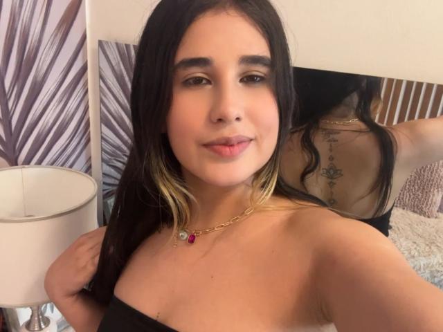 KatieDesirX, con cabello negro y rubio, vestida con un top negro sin tirantes, collar con piedras rojas y moradas, sonriendo, con un tatuaje en la espalda visible en el espejo, labios carnosos..