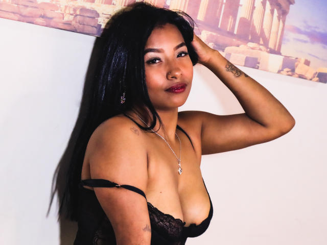 SophiaMullerr, met zwart haar, draagt zwarte kant lingerie, heeft haar hand in haar haar, toont haar borsten en een tatoeage op haar arm, en glimlacht met rode lippenstift..