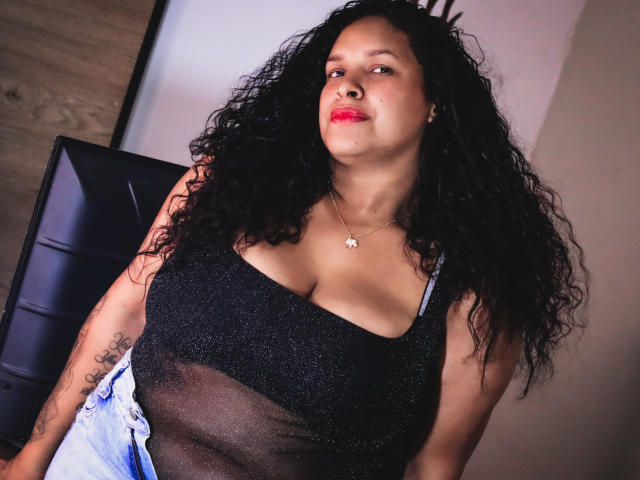 ZoehFerraro, con cabello negro rizado, lleva una blusa negra brillante que muestra sus grandes y prominentes senos, y una chaqueta azul. Tiene un tatuaje en su brazo derecho y lleva lápiz labial rojo..