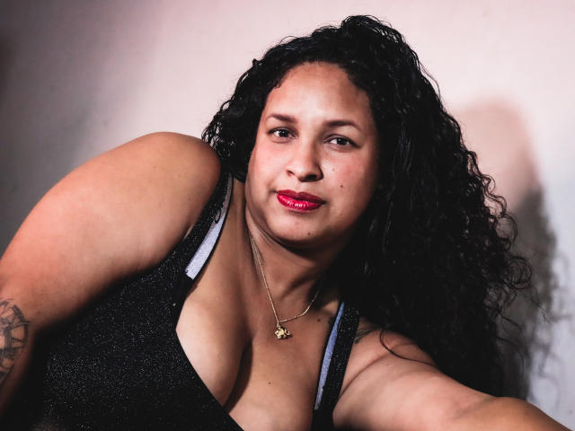 ZoehFerraro tiene el pelo negro y rizado, lápiz labial rojo y lleva una camiseta negra brillante con un collar dorado. Sus pechos grandes y redondos son visibles. Está mirando a la cámara..