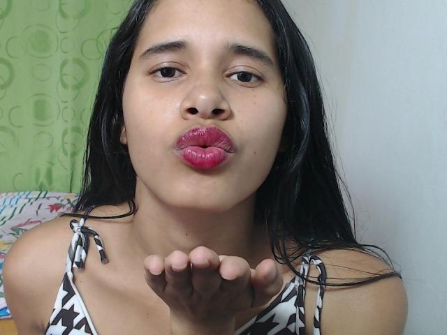 ScarlettRoss69, with wet black hair, pouty pink lips, and patterned top, blows a kiss. Her hand is raised, showing her fingers. She has a medium skin tone.
