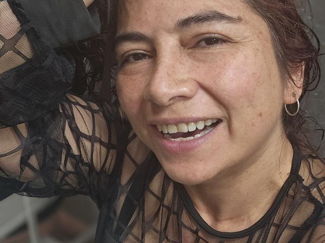 Alessia, con un sorriso felice, indossa una maglia nera in pizzo, ha delle lentiggini, orecchini a cerchio e un braccio alzato. Ha gli occhi marroni e il naso è leggermente lucido..