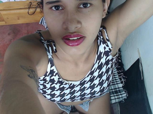 ScarlettRoss69, avec des cheveux courts et bouclés, portant un haut de bikini noir et blanc à motifs carreaux, du rouge à lèvres rose, un tatouage sur le bras gauche, regardant vers le haut, bras levé, sous-vêtements blancs visibles, tapis rose..