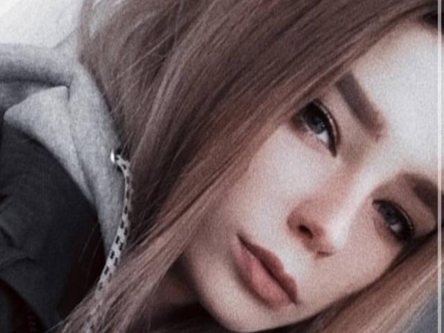 Adriene Degennaro tiene la piel clara, el cabello castaño claro y una expresión seria con los labios ligeramente entreabiertos. Lleva una sudadera gris con una cadena en el lado izquierdo..