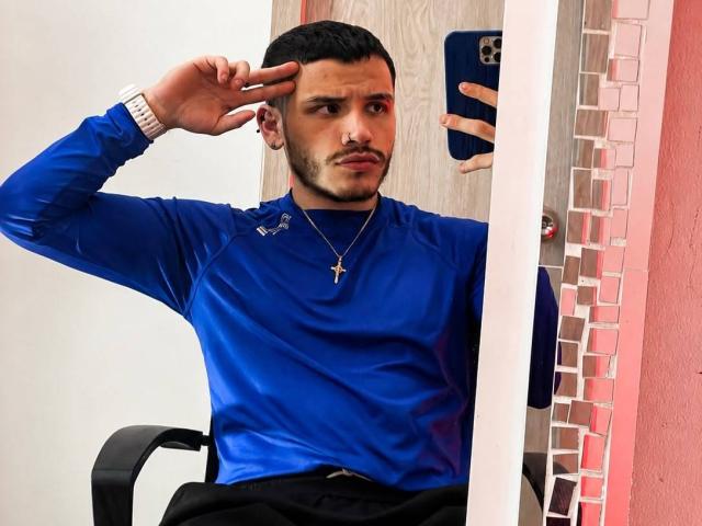 LianFerrer with a serious face, blue long-sleeve shirt, cross necklace, short black hair, making a "rock" hand sign, sitting in a chair, taking a mirror selfie.