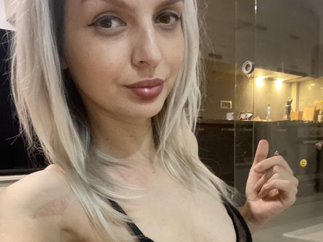 DouceAmelia has light blonde hair, wearing a black lace bra, showing off her tits, with a subtle smile, and a finger pointing upwards in a selfie.