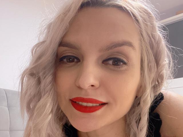 DouceAmelia's face with wavy, platinum blonde hair, red lipstick, brown eyes, and black top, showing a subtle smile and smooth skin. Boobs partially visible. Soft lighting.