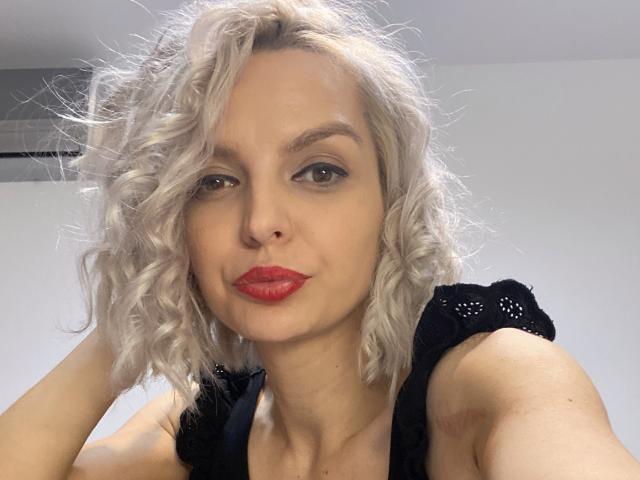 DouceAmelia with curly blonde hair, red lipstick, black lace top, smiling slightly, hands near head, pale skin, green eyes, visible nose, and a cute, confident expression.