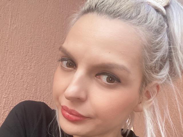 DouceAmelia's face, with gray ponytail, brown eyes, red lips, light skin, and black top, showing a slight smile, and small nose. Her tits are not visible.