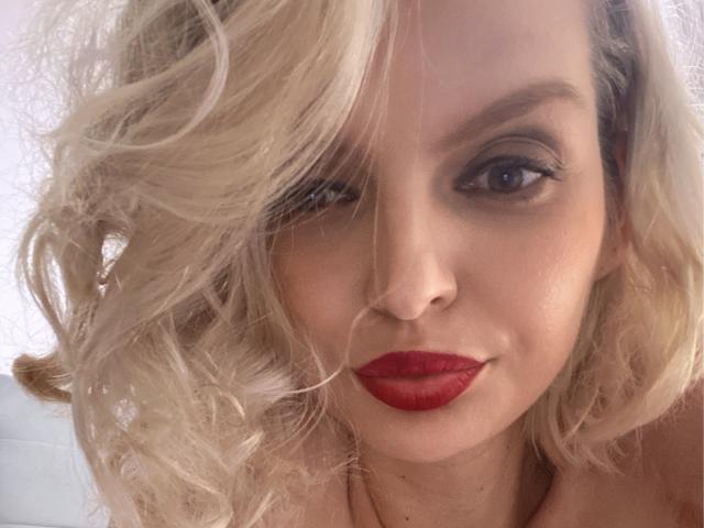 DouceAmelia has blonde, wavy hair, red lipstick, and subtle makeup, with her nose slightly tilted to the right, smiling softly at the camera. Her tits are not visible.