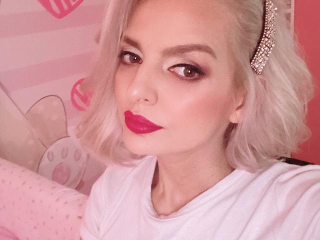 DouceAmelia with platinum blonde hair, pink gem hair clip, red lipstick, and white shirt, looking seductively at the camera. Her tits are small, and her ass is not visible.