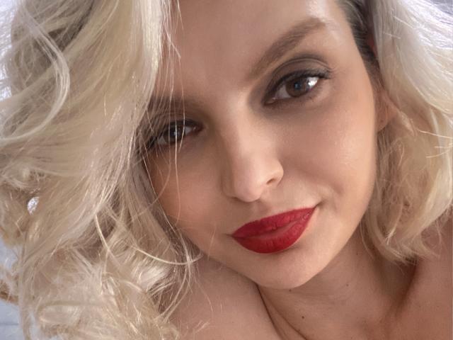 DouceAmelia's face with blonde curly hair, red lipstick, and a slight smile. She's looking directly at the camera with her brown eyes. Her tits are not visible.