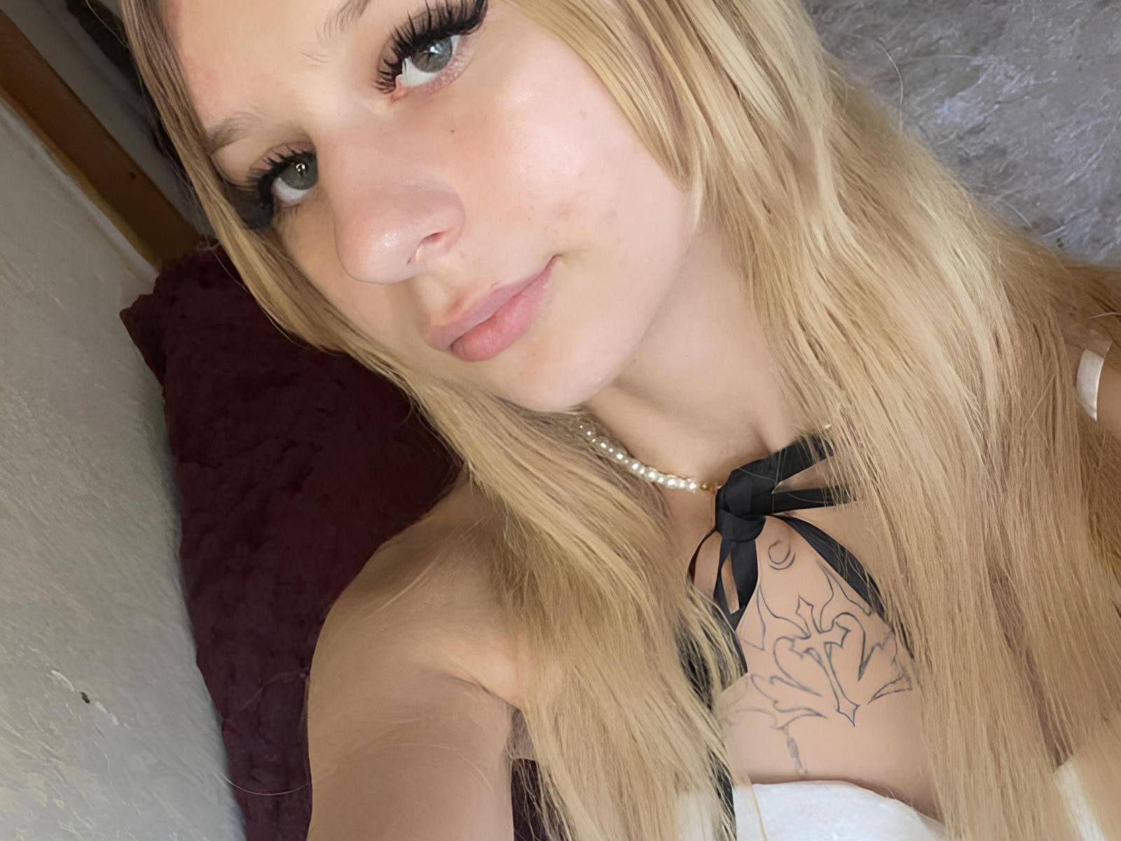 Augustina Peret, met blond haar, groene ogen en een kleine neus, draagt een parelketting en een zwarte top met een lint, en toont een bloementatoeage op haar borst en een subtiele glimlach..