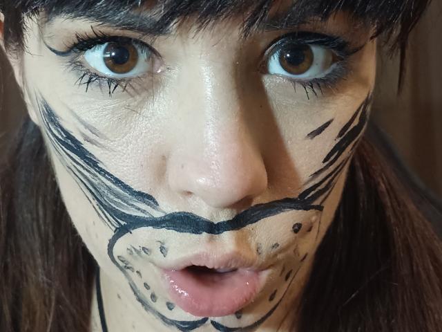 OrianaLaFrancaise's face with black painted lines resembling a mask, wide brown eyes, and slightly open pink lips. Her brown hair is partially visible. Her skin is light.
