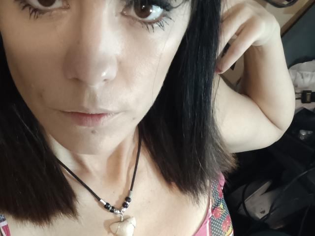OrianaLaFrancaise with shoulder-length black hair, brown eyes, and a slight pout, wearing a necklace with a white stone, touching her hair, showing a small mole on her chin.