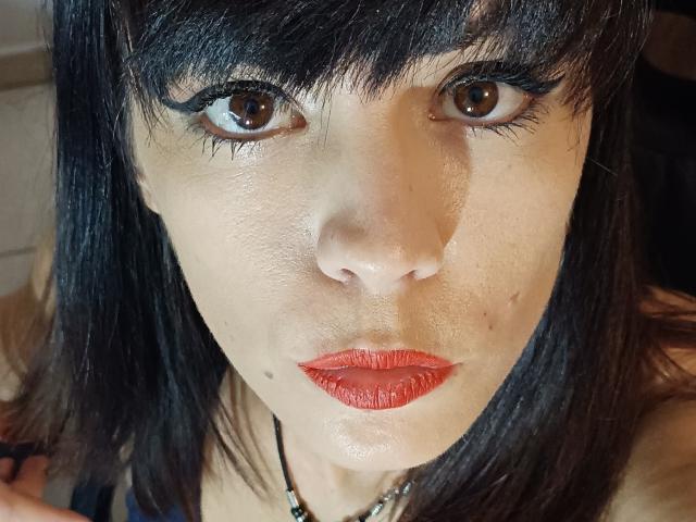 OrianaLaFrancaise has dark black hair, brown eyes, red lipstick, and a small beauty mark on her left cheek. She's looking up with a serious expression.