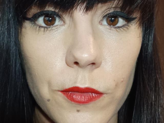 OrianaLaFrancaise has brown eyes, dark straight bangs, red lipstick, and two small moles on her right cheek. She's wearing green eyeshadow and has a slightly serious expression.