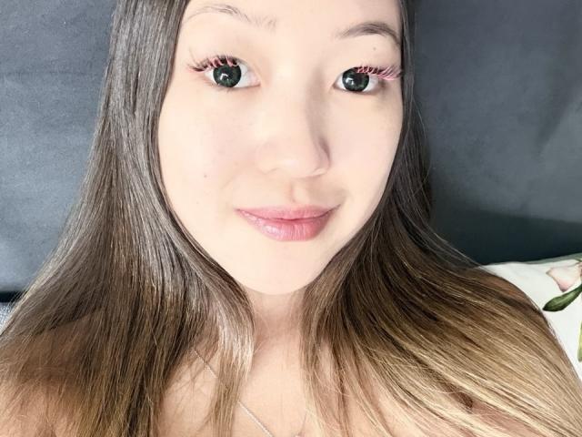 RenyLime has long brown hair, light skin, and pink lipstick, with thick red eyelashes, and a small beauty mark on her right cheek, smiling at the camera.