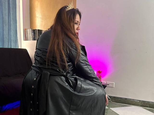 LexyDom, con cabello largo y castaño, vestida con un vestido de cuero negro y una tiara dorada, sentada en una silla, mirando hacia atrás con la boca ligeramente abierta, luz rosa a la derecha..