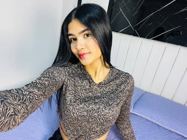 VioletteColins with long black hair, wearing a zebra-patterned top, showing her small tits, sitting on a blue bed, smiling with red lipstick, taking a selfie.