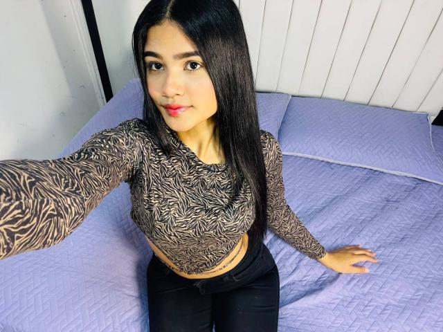 VioletteColins has long black hair, wearing a zebra-patterned crop top, sitting on a purple quilted bed, showing her tits and a small belly button tattoo.