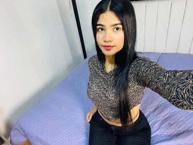 VioletteColins with long black hair, wearing a zebra-patterned crop top, showing her tits, and black pants, sitting on a purple quilted bed, taking a selfie.