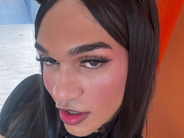 KendallKiss has shiny pink skin, thick black eyebrows, long eyelashes, and pink lipstick. She's looking up with dark hair, wearing a black top. Her nose is slightly shiny.