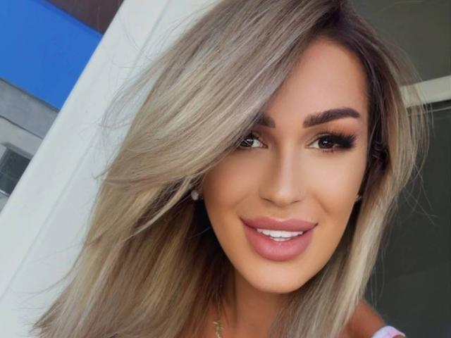 NicolSNaya has blonde hair, brown eyes, thick eyebrows, long eyelashes, and pink lipstick. She's smiling with a cute, sexy face. Her tits aren't visible in this shot.