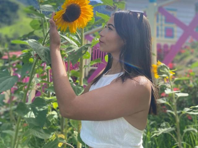GraceLunaWallace mit gebräunter Haut, schwarzen Haaren, einem weißen Top ohne Ärmel, die eine hohe Sonnenblume berührt. Sonnenlicht, grüne Blätter, ein rosa und gelber Hintergrund. Brüste sind sichtbar, Seitenansicht, Fokus auf der Blume..