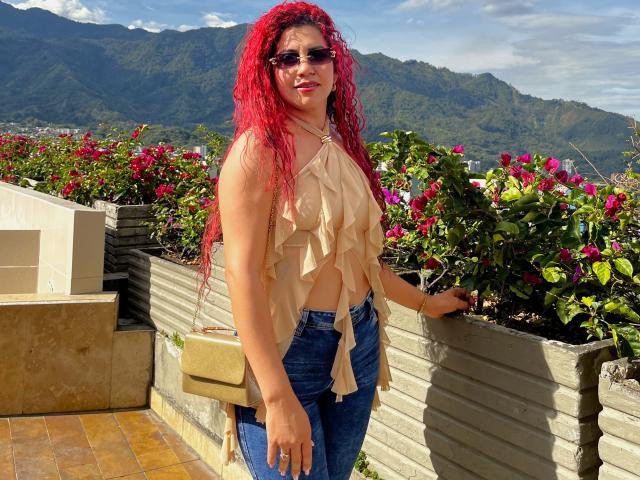 AntonelaRiveiro with bright red curly hair, sunglasses, beige ruffled top, blue jeans, and tan crossbody bag, standing on a sunlit terrace with pink flowers and mountains in background.