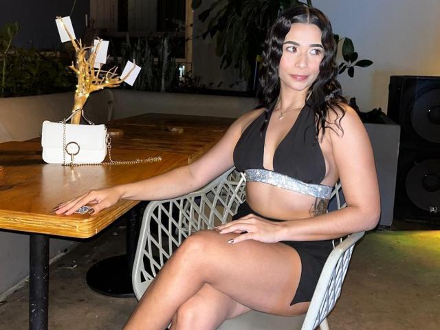 GraceLunaWallace mit dunklen, welligen Haaren, schwarzem Bikini-Oberteil, das ihre Brüste zeigt, schwarzen Shorts, sitzend auf einem weißen Stuhl, Hand auf einem Holztisch, weiße Handtasche, goldener Baum, Lautsprecher im Hintergrund..