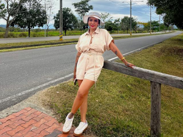 JessicaFiore, standing by a wooden fence, wearing a beige button-up romper, white sneakers, and a white sun hat, with a tattoo on her left arm and a smile.