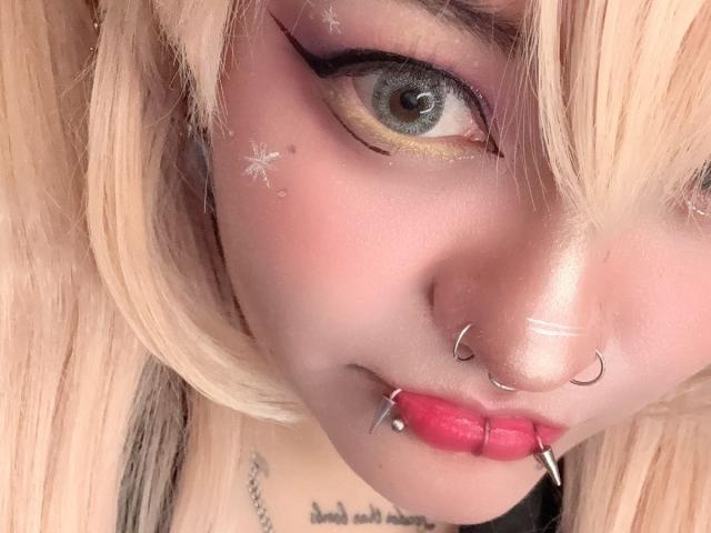 LuckyLeahh's face with blonde hair, blue eye, pink eyeliner, nose piercings, lip piercings, pink lipstick, tattoos, and sparkly makeup. Boob tattoo barely visible under chin.