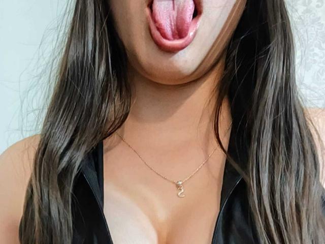 AnnFisher with her tongue out, showing pink tongue and slightly parted lips, wearing a black top with a gold necklace, exposing her tits. Her brown hair is down.