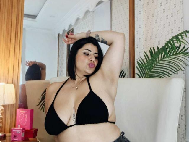 AngeleRouxo with black bob, puffy lips, black bikini top, showing big tits, arm raised, tattoo on arm, in living room, white couch, green plant in background.