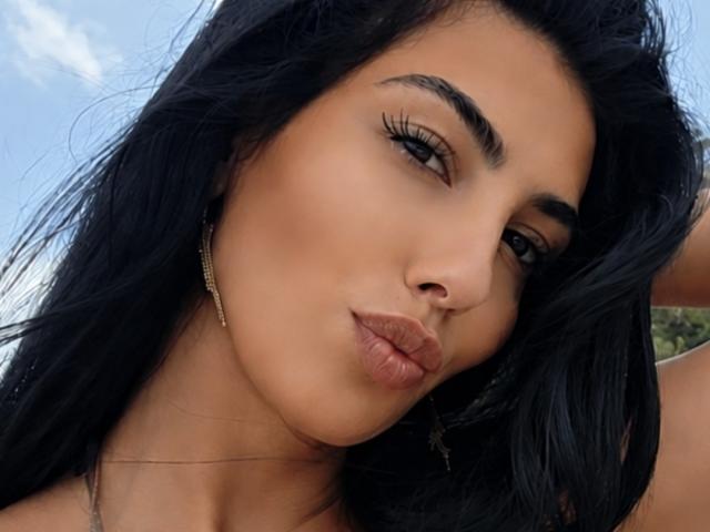 BrunetteBabe69 has pouty lips, dark eyebrows, long black eyelashes, and is making a pensive face with her head slightly tilted. She has tan skin and is wearing gold earrings.
