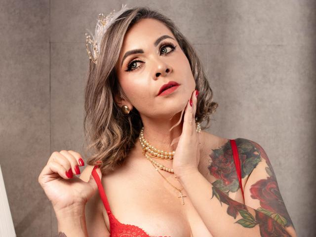 VictoriiaGreys with red bra, pearl necklace, and crown, holding strap, showing tattooed arm with roses, and red nails, looking up with hand on cheek, and cross necklace.