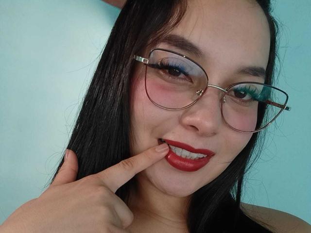MelaniCoox with long black hair, wearing red lipstick and glasses, biting her finger with a cute smile. Her blue eyeshadow and pink cheeks are visible.
