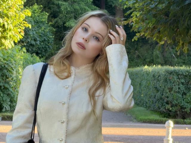 StellaMelow has blonde hair, blue eyes, and a light button-up shirt with a black strap. She's touching her head, looking slightly confused, with a tree in the background.