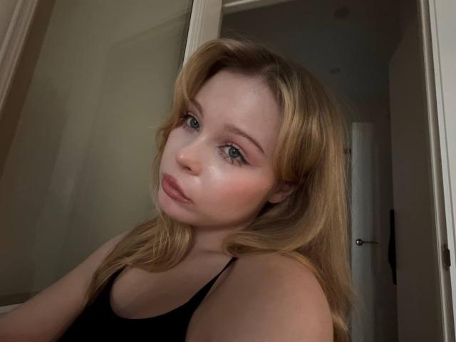 StellaMelow with blonde hair, blue eyes, and pinkish skin, wearing a black tank top, looking slightly to the side with a neutral expression. Boobs visible, soft lighting.