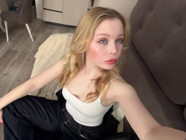 StellaMelow with blonde wavy hair, blue eyes, pink lips, white tank top, black jeans, sitting on floor, looking at camera, holding arm out, gray sofa in background.