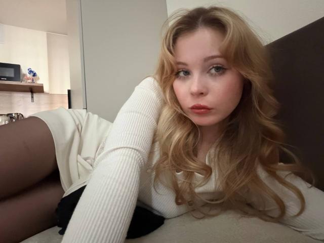 StellaMelow with long blonde hair, lying on a bed, wearing a white ribbed top, black shorts, and black tights, looking at the camera with pouty red lips.