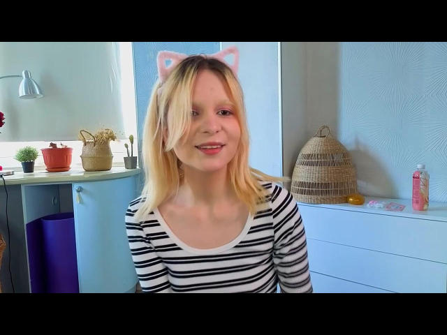 AnnikaFradette with blonde hair, cat ears, and a striped shirt, smiling with pink lips, showing a cute, playful expression, sitting in a bright room.