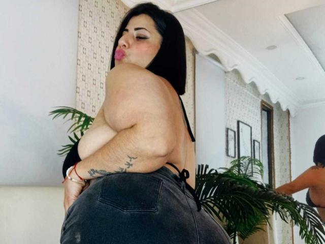 AngeleRouxo with large ass, black hair, and tattoos, wearing a black halter top and dark jeans, pouting with her back to the camera, in a modern room.