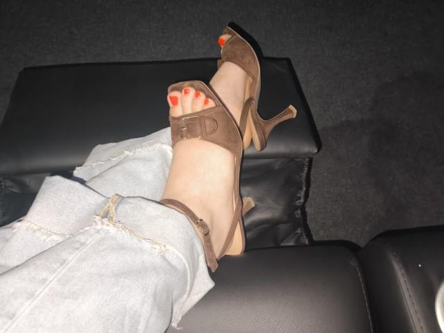 Dreia's foot with red-painted toes in brown high-heeled sandals, crossed over her denim-covered ankle, resting on a black leather seat.