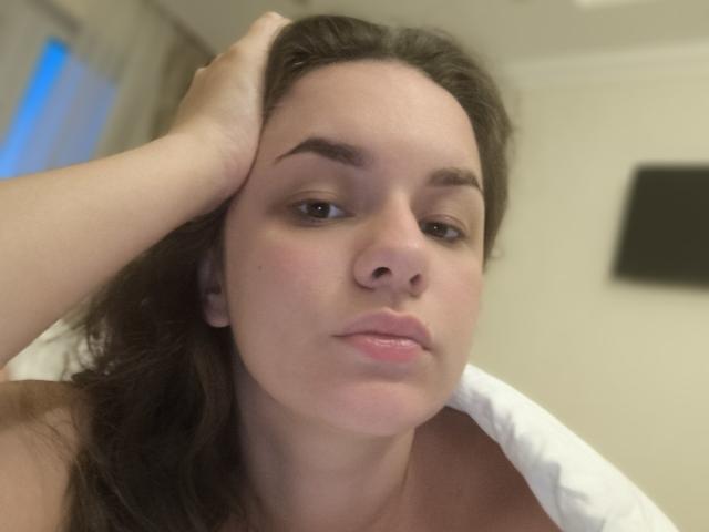 Dreia met bruin haar, licht open lippen en hand in haar, kijkt naar de camera en laat zachtroze tepels zien onder een wit kussen. Vage achtergrond. Informele, licht onscherpe foto..